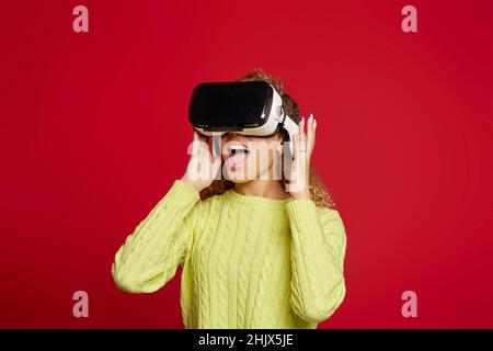 Une femme afro-américaine stupéfait dans des lunettes de la réalité virtuelle explorant le cyberespace sur fond rouge en studio Banque D'Images