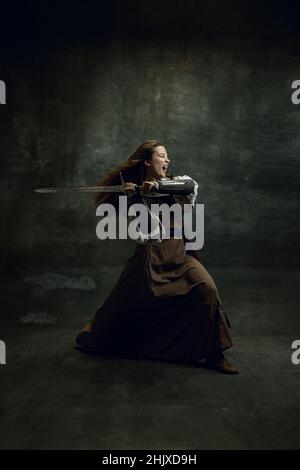 Portrait dynamique de seriuos belle femme à l'image du guerrier médiéval ou chevalier avec sale visage blessé avec grande épée isolée sur vintage sombre Banque D'Images