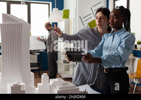 Femme architecte pointant un crayon sur le modèle architectural de gratte-ciel debout à côté de l'ingénieur de collègue tenant une tablette dans un bureau moderne.Architectes faisant le travail d'équipe regardant la maquette de projet résidentiel. Banque D'Images