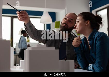 L'équipe des architectes pointe le stylo vers le gratte-ciel pour discuter de la conception de l'architecture dans un bureau moderne.Les architectes conbralisent les idées en collaborant sur le modèle en mousse blanche. Banque D'Images