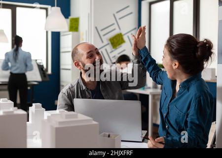 Deux architectes de haut fiving assis au bureau dans un bureau moderne architectural avec ordinateur portable et maquette en mousse de bâtiments résidentiels.Des collègues souriants et heureux qui célèbrent le projet d'équipe. Banque D'Images