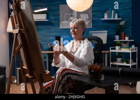 Artiste âgé assis devant un chevalet avec smartphone de navigation en toile pour l'inspiration.Fier créateur d'art senior affichant un croquis de charbon de bois de chef-d'œuvre sur les médias sociaux à l'aide d'un appareil numérique. Banque D'Images