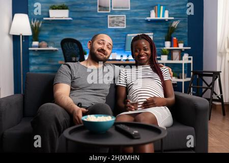 Couple multiracial appréciant la télévision et attendant bébé à la maison tout en se relaxant sur le canapé.Les gens adorables se sentent heureux de regarder une comédie décontractée ensemble à la télévision dans le salon en attendant que bébé soit né. Banque D'Images