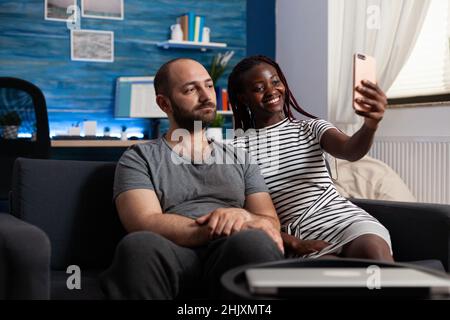 Souriante femme afro-américaine prenant le selfie à l'aide d'un smartphone moderne tout en ennuyé homme caucasien posant.Divers couples prenant des selfies qui se tardent d'un appareil intelligent moderne tout en étant assis sur un canapé à la maison Banque D'Images