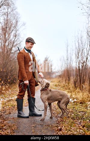 Chasseur en costume marron avec chien fort de la race Weimaraner sur le paysage du début de l'automne, se préparer à la chasse, l'entraînement masculin du chien, donnant le comman Banque D'Images