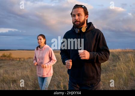 Jeune homme concentré et femme courant ensemble pendant le coucher du soleil, jogging dans la campagne. Mâle caucasien barbu actif et en forme, partenaire femelle dans le dos Banque D'Images