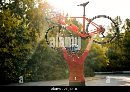 Regardez le Sporty man dans des vêtements de sport, un casque de protection et des lunettes transportant le vélo levant après une compétition réussie, en posant à la caméra avec humour Banque D'Images