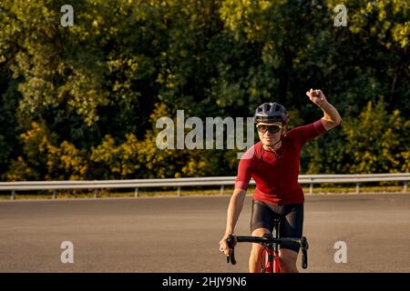 Jeune cycliste caucasien européen en casque noir, lunettes de protection et rouge actif sportive porter dynamiquement vélo, heureux après la compétition. Banque D'Images