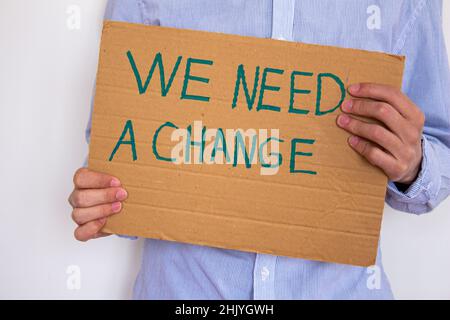 Homme tenant un carton avec une inscription « We Need a change » sur fond de mur gris.Il peut protester contre le réchauffement de la planète ou la pollution de l'environnement. Banque D'Images