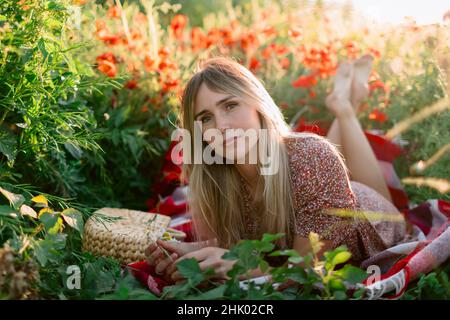 Portrait d'une femme en plein air sur un motif écossais avec des fleurs de pavot au coucher du soleil.Femme en robe et sac à main avec fleurs de champ Banque D'Images