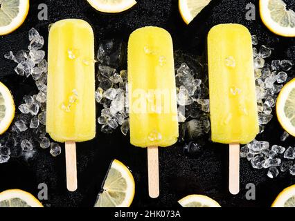 Trois pop-cicles de citron sur fond noir avec de la glace et des tranches de citron.Composition de la pose à plat. Banque D'Images