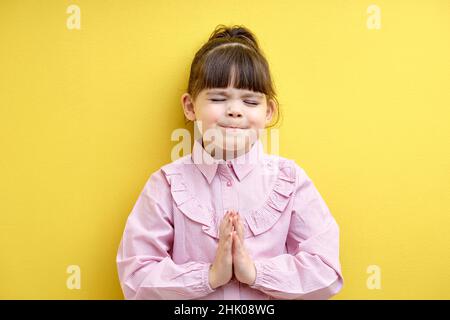 petite fille mendiant à acheter, demandez quelque chose. enfant doux dans un chemisier rose garder les mains ensemble, isolé sur fond jaune, debout avec les yeux fermés Banque D'Images