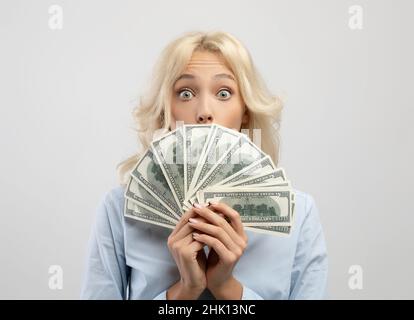 Femme d'affaires choquée couvrant son visage avec de l'argent, se cachant face derrière un fan de dollar dans ses mains, fond clair Banque D'Images