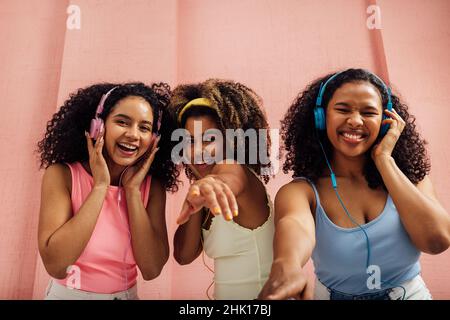 Trois femmes riant avec des cheveux bouclés dansant ensemble et regardant l'appareil photo, en portant des écouteurs Banque D'Images