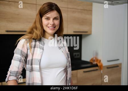 Portrait d'une belle femme enceinte européenne debout dans la cuisine et souriante regardant l'appareil photo Banque D'Images