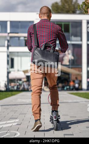 Jeune homme dans un pantalon décontracté et une chemise à cheval sur son scooter électrique, voir la forme derrière, les bâtiments à l'arrière-plan Banque D'Images