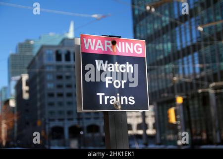 Avertissement attention aux trains, panneau noir et rouge, avec des bâtiments à l'arrière et reflétant le soleil Banque D'Images