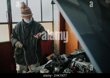 Vieil homme avec masque de protection dans le garage réparer sa vieille voiture. Banque D'Images