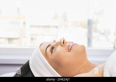 Une femme heureuse souriant étant dans la clinique de beauté avec une serviette de bandage blanche sur son front se reposant après la procédure de massage facial. Banque D'Images