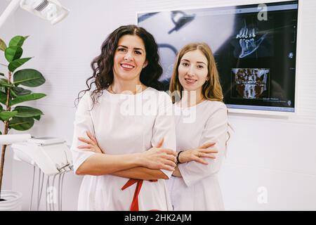 Deux médecins dentistes femme heureux de regarder la caméra portant l'uniforme blanc debout dans la clinique de bureau avec la dent instantané à l'ordinateur plus Banque D'Images