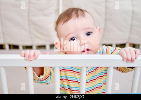 Un bébé triste dans un berceau, tenant sur les mains courantes.Enfant drôle au lit, six mois Banque D'Images