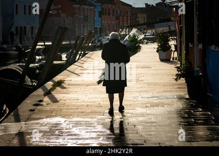 Burano, Italie - 26 octobre 2021: Vieille femme avec des fleurs marchant à Burano près du canal, jour ensoleillé en automne Banque D'Images