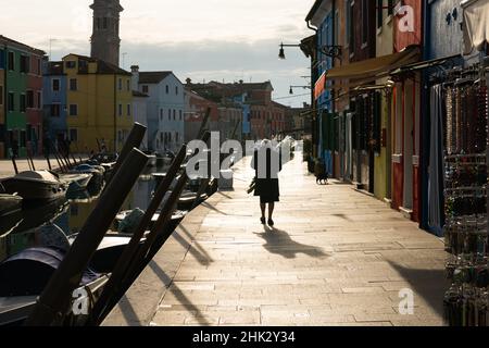 Burano, Italie - 26 octobre 2021: Vieille femme avec des fleurs marchant à Burano près du canal, jour ensoleillé en automne Banque D'Images