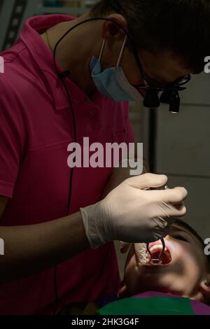 Le dentiste pédiatrique traite les dents de l'enfant, pose d'un barrage en caoutchouc pour l'enfant, lumière locale sur le visage du patient. Banque D'Images