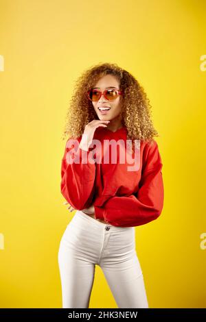 Jeune femme afro-américaine confiante portant un élégant sweat à capuche rouge et des lunettes sur fond jaune en studio Banque D'Images