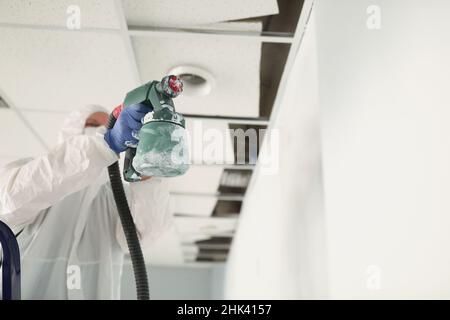 Ouvrier de construction mâle dans des combinaisons de protection peinture mur blanc avec pistolet de pulvérisation de gros plan Banque D'Images