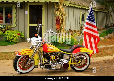 Harley Davison orange et jaune avec un drapeau américain monté à l'arrière, garé devant un magasin décoré pour l'automne. Banque D'Images