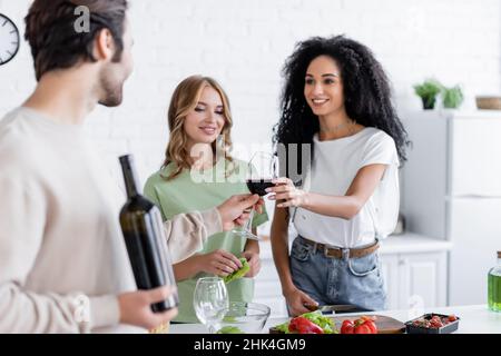 homme flou donnant un verre de vin à une femme afro-américaine joyeuse près d'une amie blonde dans la cuisine Banque D'Images