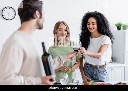 homme flou donnant un verre de vin à une femme afro-américaine heureuse près d'une amie blonde dans la cuisine Banque D'Images