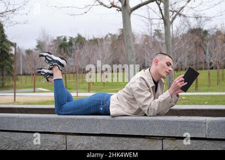 Un jeune homme portant du maquillage pose sur un mur avec les jambes croisées lisant un livre. Banque D'Images