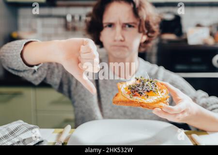 adolescente avec cheveux bruns, émotion insatisfaite sur le visage, montrant le pouce vers le bas geste de désapprobation, condamnant l'oeuf et le sandwich de microgreens, Banque D'Images