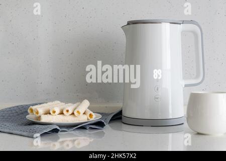 Sur le comptoir de cuisine se trouvent des rouleaux de galette sur une assiette avec une bouilloire électrique blanche et une tasse.Pause café. Banque D'Images