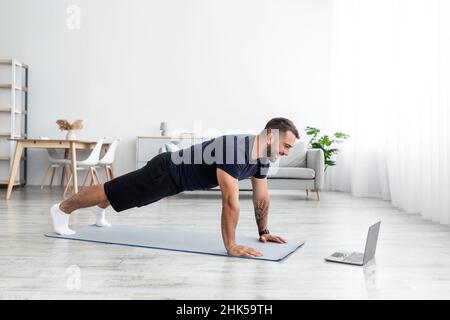 Joyeux millénaire musclan caucasien mâle faisant de la planche et regardant la montre d'ordinateur en ligne leçon dans le salon Banque D'Images