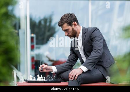 Homme d'affaires banquier jouant aux échecs assis sur le banc près du bureau, pensif faisant un mouvement stratégique Banque D'Images