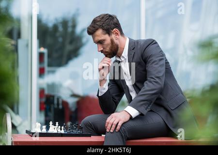 Homme d'affaires banquier jouant aux échecs assis sur le banc près du bureau, pensif faisant un mouvement stratégique Banque D'Images