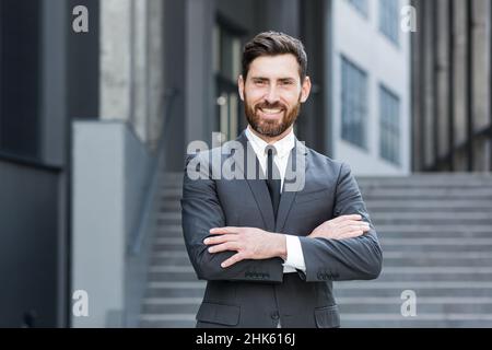 Homme d'affaires prospère et joyeux regardant l'appareil photo et souriant, portrait avec les bras croisés près du bureau Banque D'Images