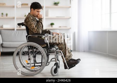 Mécontent jeune homme regardant la caméra avec un regard triste. Il est assis dans une chaise invalide portant un uniforme militaire. Isolé sur fond gris. Copier l'espace Banque D'Images