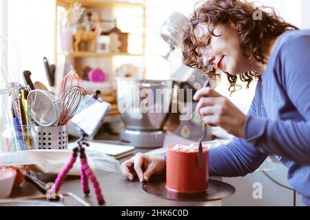 Jeune femme à la maison enseignant un cours de boulangerie en ligne.Elle utilise un téléphone portable sur un trépied.Blogging de nourriture. Banque D'Images