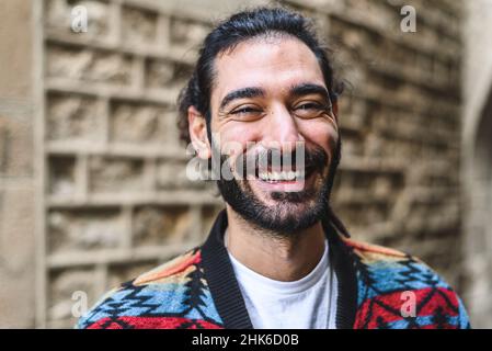 Joyeux hispanique dans une veste colorée avec barbe et queue de cheval souriant et regardant l'appareil photo. Tout en se tenant sur un fond flou de mur de briques dans la rue Banque D'Images
