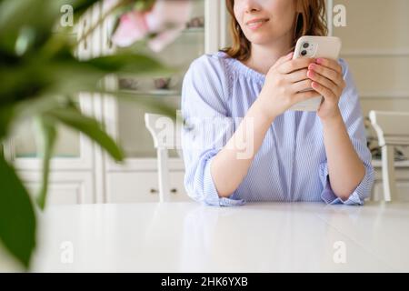 Une jeune femme avec un téléphone mobile moderne est assise à une table blanche en souriant derrière des fleurs floues au premier plan dans la pièce à la maison vue rapprochée Banque D'Images