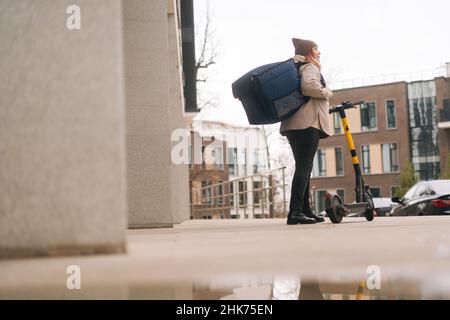 Vue arrière à angle bas de la livraison de nourriture de coursier femelle avec grand sac à dos thermique debout avec scooter électrique dans la rue de ville. Banque D'Images