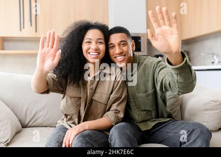 Écran vidéo avec un couple multiracial joyeux, regardant la caméra et en agitant.Webcam vue de jeune biracial souriant femme et homme assis sur le canapé et salutation, concept de réunion virtuelle Banque D'Images