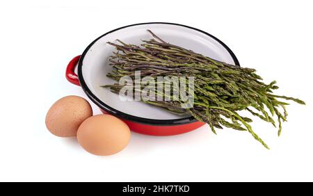 Bouquet d'asperges sauvages crues dans un vieux pot en émail rouge sur fond blanc Banque D'Images