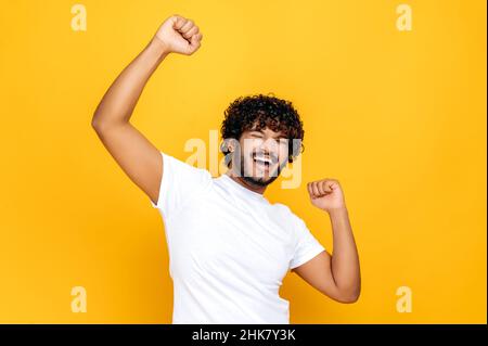 L'homme indien, chanceux et agacé, dansant, gestinant avec des poings, reçoit des bénéfices, heureux de gagner une loterie, debout sur fond orange isolé.Bonne nouvelle, succès et concept de bonheur Banque D'Images