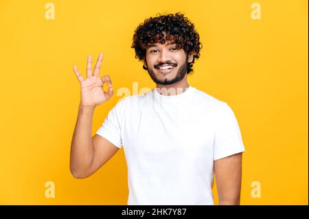 Un sympathique garçon indien ou arabe à poil dur dans un t-shirt blanc, montrant le signe ok tout en se tenant sur un arrière-plan orange isolé, regarde l'appareil photo, souriant amical Banque D'Images