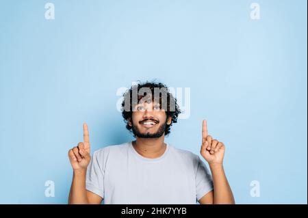Jeune homme indien ou arabe positif en t-shirt de base, surpris regarde vers le haut et pointe les doigts vers le haut, à l'espace vide, se tient sur un arrière-plan isolé de couleur orange.Maquette, concept d'espace de copie Banque D'Images